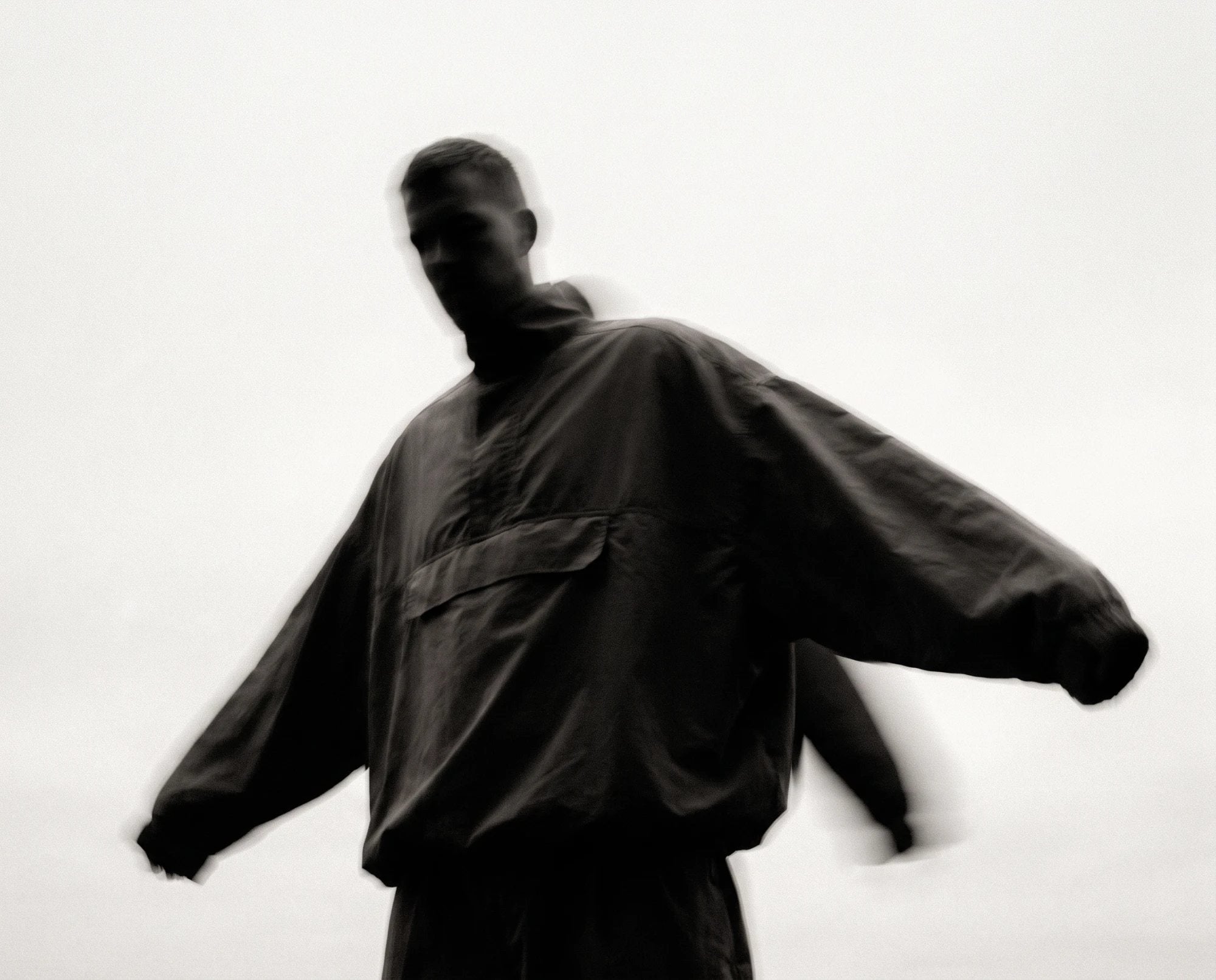 A man in a dark oversized anorak, blurred in motion against a pale sky